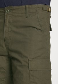 Pantalones cargo verde oliva hechos de un tejido ligero, con textura a cuadros, cierre frontal con botón y detalle de bolsillos laterales.