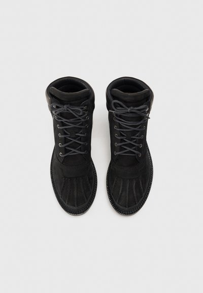Bottes noires mi-hautes avec une tige en daim, une pointe ronde et des coutures texturées. Lacets gris et col rembourré pour le confort.