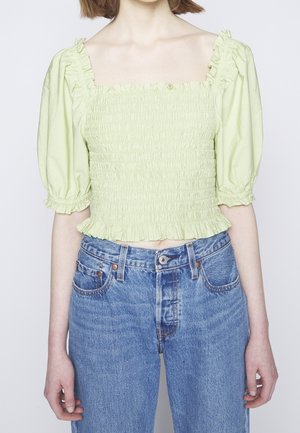 Jasnozielony marszczony crop top z bufkami, zestawiony z wysokim stanem niebieskich jeansów na osobie z krótkimi włosami.