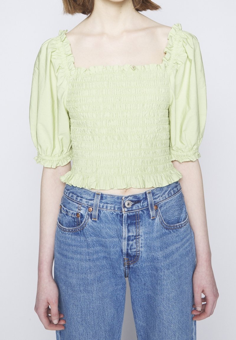 Crop top smocké vert clair avec manches bouffantes, associé à un jean en denim bleu taille haute sur une personne aux cheveux courts.