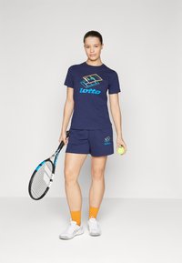 Dunkelblaues Sport-T-Shirt und Shorts mit Lotto-Logo. Mit lebhaftem Grafikdruck und passenden Akzenten, kombiniert mit einem Tennisschläger und einem Ball.