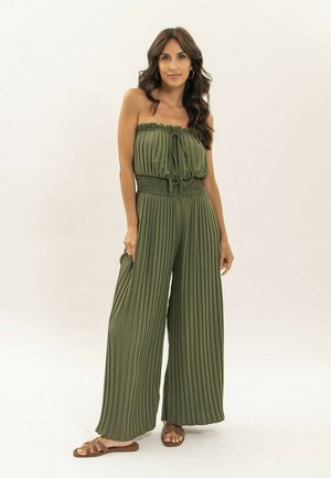 Femme aux longs cheveux ondulés portant une combinaison à bretelles olive plissée et des sandales marron, debout devant un fond clair uni.