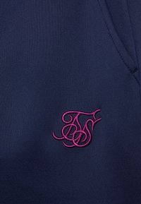 Marinblått tyg med en slät yta har detaljer av en rosa broderad logotyp. Designen inkluderar en subtil fickkontur.