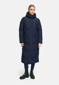 Lang marineblauwe pufferjas met een chevron-quiltpatroon, hoge kraag, veilige capuchon en zijzakken, gecombineerd met zwarte enkelboots.