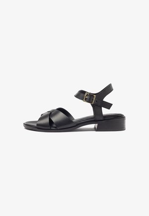 Sandale en cuir noir avec un petit talon bloc, des brides croisées à l'avant et une bride de cheville avec boucle dorée sur fond blanc.