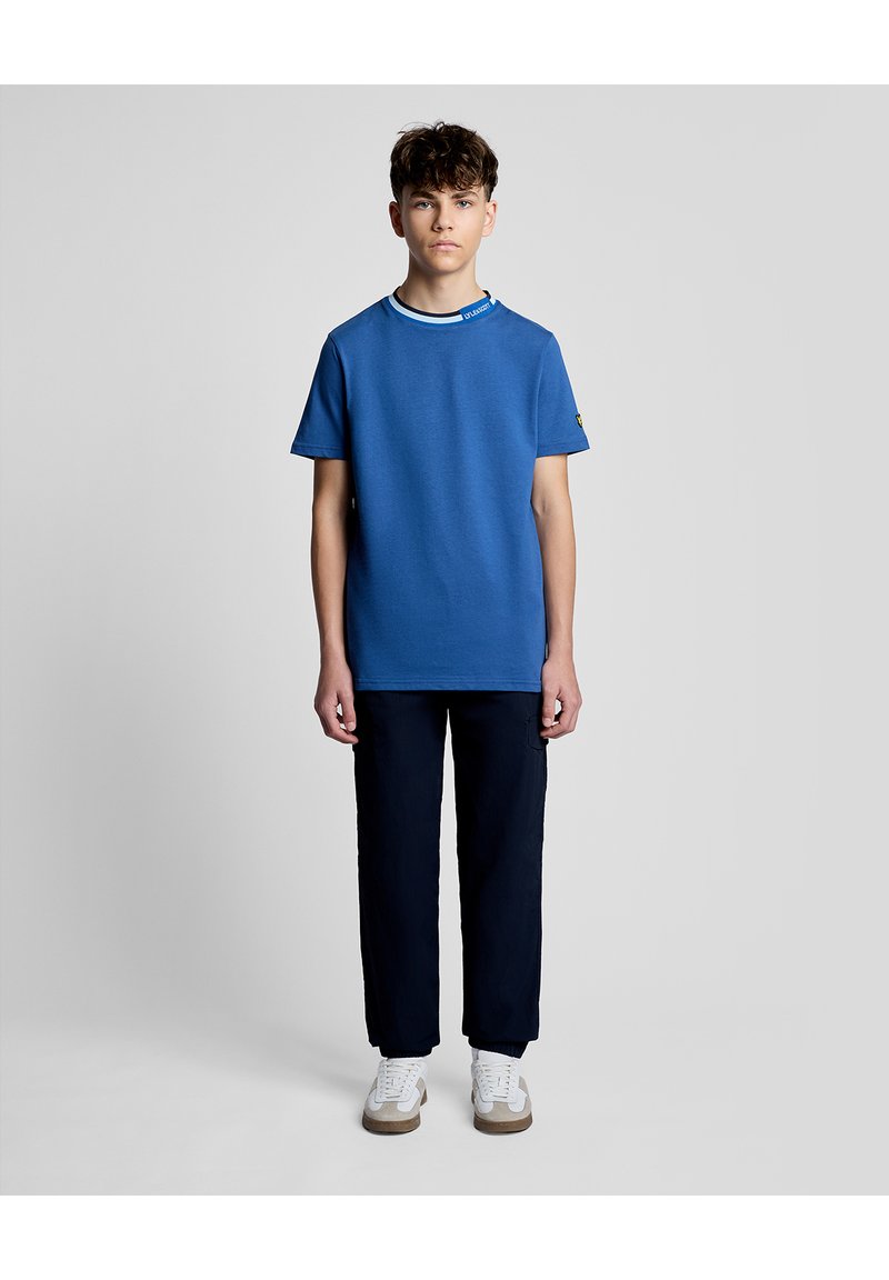Adolescent debout, portant un t-shirt bleu à manches courtes, un pantalon foncé et des baskets blanches, sur un fond uni gris clair.