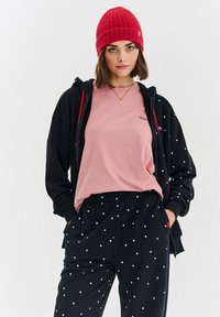 Czarny rozpinany hoodie z czerwonymi akcentami na różowej koszulce. Czarne dresy w białe kropki z elastycznym paskiem w talii. Czerwona dziana czapka.