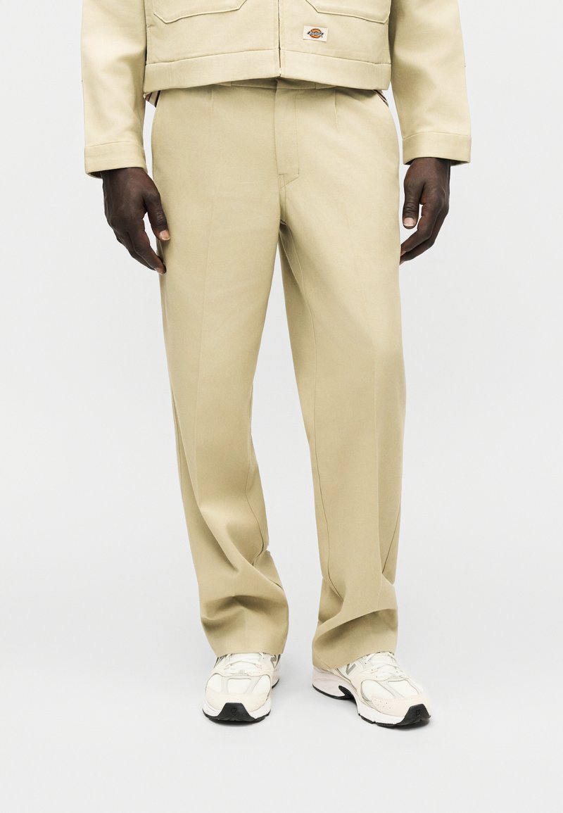 Pantalon beige à jambes droites porté avec des baskets blanches et une veste beige, vue de face de la taille jusqu'aux pieds.
