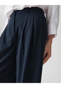 Marineblauwe op maat gemaakte broeken met plooien, een zachte textuur, zijzakken en een aansluitende tailleband. Gepaard met een wit overhemd met lange mouwen.