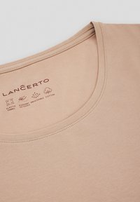 Beżowa bawełniana koszulka z okrągłym dekoltem, o gładkiej fakturze. Na materiale nadrukowane instrukcje pielęgnacji i nazwa marki w ciemnoczerwonym kolorze.