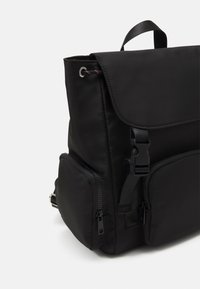 Sac à dos en tissu noir avec fermeture à rabat, sangle réglable et deux poches latérales zippées, doté d'une texture lisse et d'un design minimaliste.