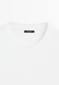 Gros plan sur un t-shirt blanc uni à col rond avec une étiquette noire Massimo Dutti cousue à l’intérieur du col.