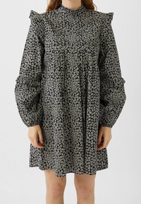 Nainen, jolla on päällään löysä musta mekko, jossa on pieniä valkoisia kukkakuoseja, pitkät puffihihat, rypytyksillä olkapäillä ja kyynärpäiden ympärillä.