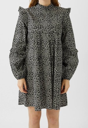 Donna che indossa un vestito nero largo con una piccola stampa floreale bianca, maniche lunghe a sbuffo, dettagli a volants sulle spalle e intorno ai gomiti.