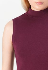 Närbild av en person som bär en ärmlös, höghalset shirt i vinröd färg, med ljus hy och blont hår synligt.