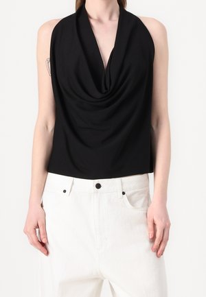 Femme portant un haut drapé noir sans manches à col noué, associé à un pantalon blanc taille haute, debout les bras détendus.