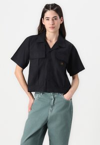 Camisa preta cropped com botões, dois bolsos frontais, mangas curtas e um tecido texturizado. Usada com calças verdes claras de cintura alta.