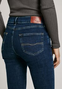 Tmavomodré džínsy z denimu s koženou nášivkou na zadnom páse, obsahujúce dve zadné vrecká s kontrastným prešívaním.