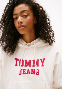 Felpa bianca realizzata in tessuto morbido, con un grande logo rosso "TOMMY JEANS". Presenta un cappuccio con coulisse e una tasca frontale.