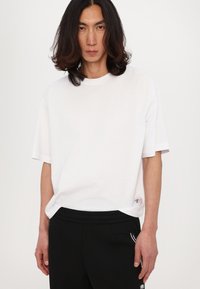 T-shirt bianco a maniche corte realizzato in cotone. Presenta un colletto rotondo e una piccola toppa con logo all'orlo. Abbinato a pantaloni neri.