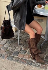 Brązowe, zamszowe kozaki za kolano z frędzlami, z zaostrzonym noskiem. W tle ciemna skórzana kurtka, czarna spódnica oraz fakturowana brukowana nawierzchnia.