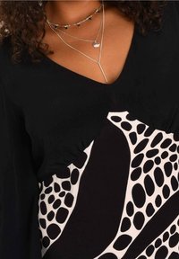 Svart blus med v-ringning, med vita abstrakta mönster och texturerade volanger längs axlarna. Lager av silverhalsband ger detaljer.