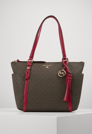 Brązowa torebka Michael Kors z czerwonymi skórzanymi uchwytami, bocznymi kieszeniami, złotym brelokiem z logo i detalem z chwostem na jednolitym tle.