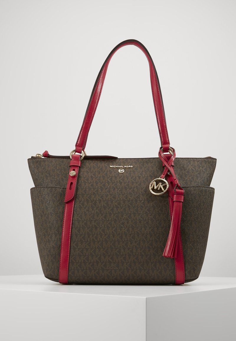 Sac à main Michael Kors marron avec poignées en cuir rouges, poches latérales, breloque logo dorée et détail pompon sur fond uni.