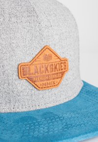 Szürke szövet sapka türkiz peremmel. Jellemzője egy barna bőrkészlet, amelyen a "BLACKSKIES PREMIUM SUPPLIES" felirat található, valamint texturált részletek.