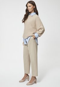 Beżowy sweter robiony na drutach, jasnoniebieska koszula z kołnierzykiem pod spodem, beżowe spodnie zwężane i beżowe buty na słupku. Gładkie białe tło.