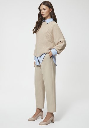 Beżowy sweter robiony na drutach, jasnoniebieska koszula z kołnierzykiem pod spodem, beżowe spodnie zwężane i beżowe buty na słupku. Gładkie białe tło.