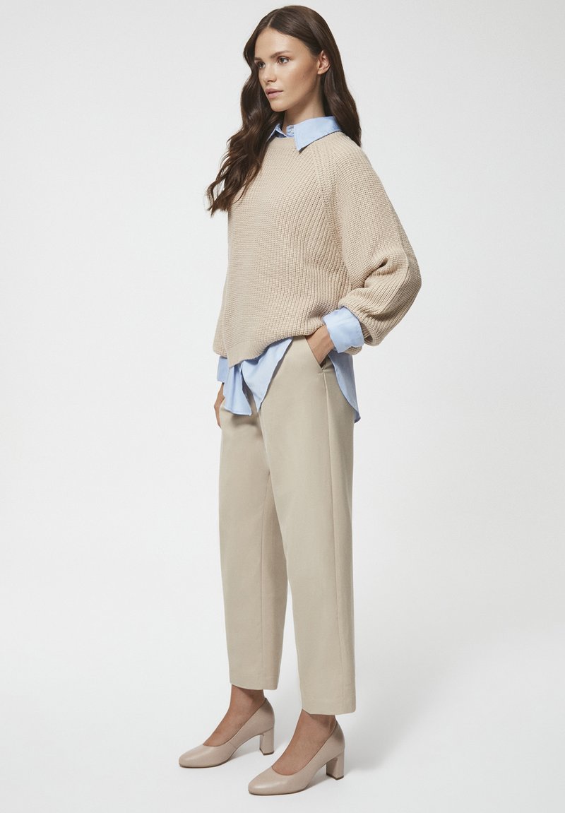 Beżowy sweter robiony na drutach, jasnoniebieska koszula z kołnierzykiem pod spodem, beżowe spodnie zwężane i beżowe buty na słupku. Gładkie białe tło.
