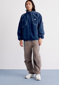 Person wearing a navy fleece jacket with zipper, loose beige pants with side stripes, and white sneakers standing on a blue floor.