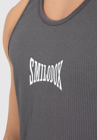 Grijs geribbeld tanktop met een ronde hals en een gebogen witte "SMILODOX" logo. De stof lijkt glad met een subtiele textuur.