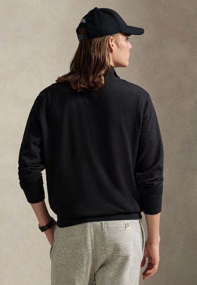 Person mit langen Haaren, die eine schwarze Kappe, ein schwarzes Sweatshirt und hellgraue Jogginghose trägt, mit dem Rücken zu einem neutralen Hintergrund.