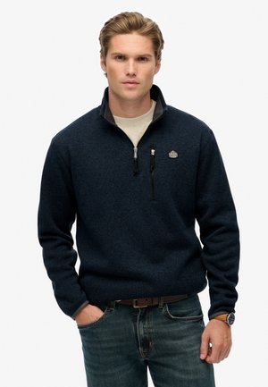Sudadera azul marino con cremallera hecha de tejido de punto texturizado, que presenta un cuello alto, un bolsillo en el pecho y un detalle de logo en el pecho.