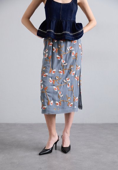 Top peplum de denim combinado con una falda gris hasta la rodilla, adornada con bordados florales en naranja y blanco. Zapatos de tacón alto negros y puntiagudos.