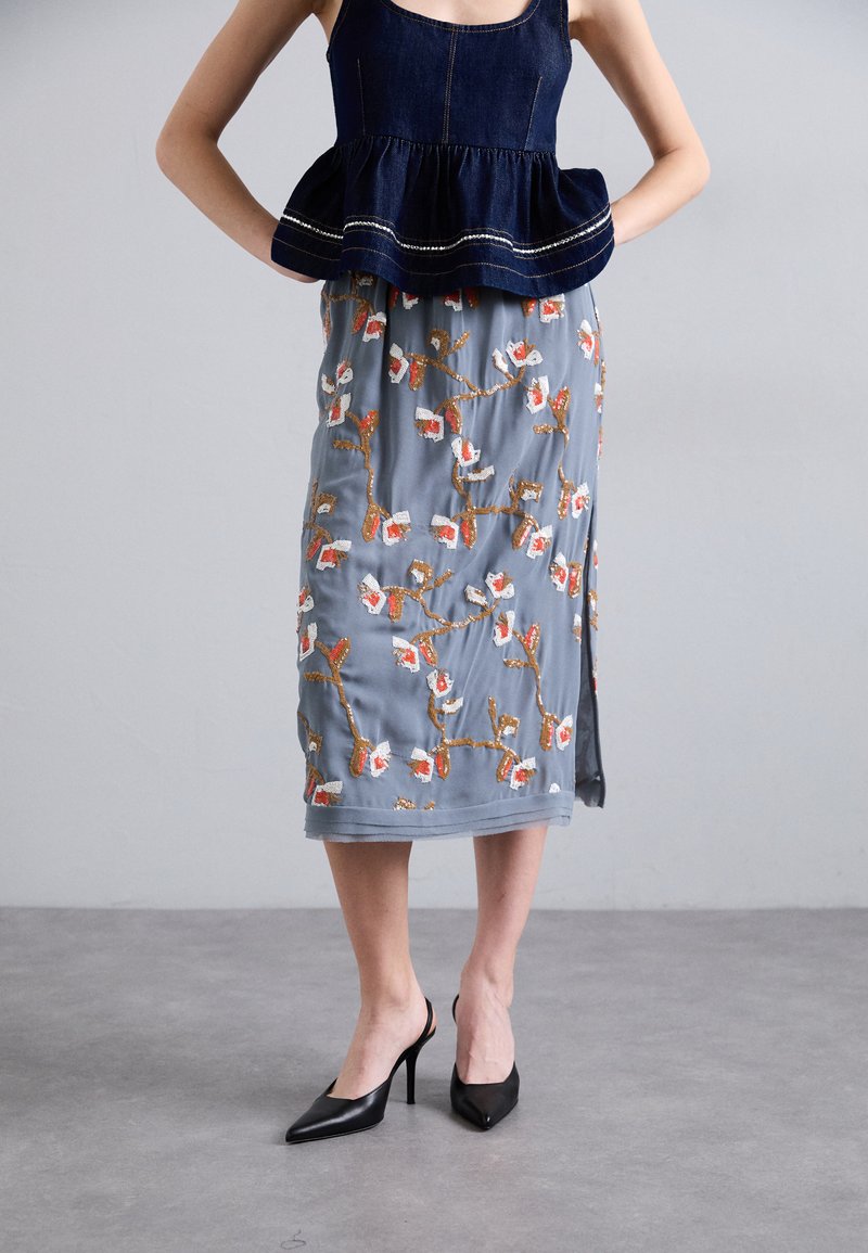 Τζιν μπλούζα peplum συνδυασμένη με μια γκρίζα φούστα μήκους γόνατων που έχει πορτοκαλί και λευκή κεντημένη ανθοφορία. Μαύρες ψηλοτάκουνες γόβες.