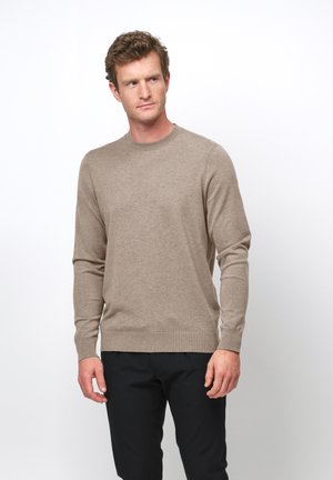 Mann in beigem Rundhalspullover und schwarzer Hose steht vor weißem Hintergrund und schaut leicht zur Seite.