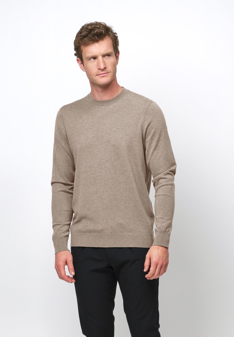 Hombre con suéter beige de cuello redondo y pantalones negros, de pie contra un fondo blanco, mirando ligeramente hacia un lado.