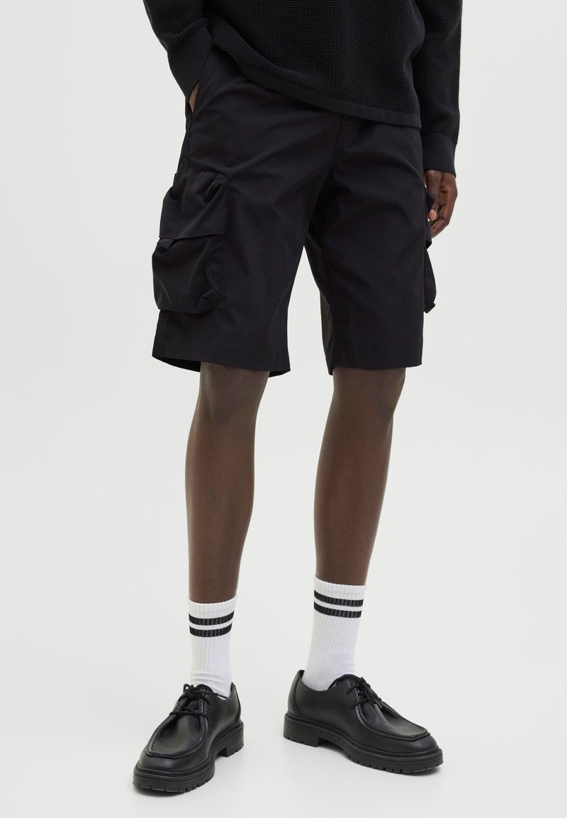Personne portant un short cargo noir, des chaussettes blanches avec deux bandes noires et des chaussures noires à lacets sur un fond clair uni.