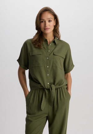 Olivnozielony kombinezon z krótkim rękawem, z zapięciem na guziki, dwiema kieszeniami na piersi i wiązanym detalem w talii. Wykonany z lekkiego materiału.