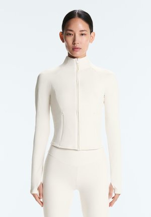 Donna che indossa un top crema aderente con zip e maniche lunghe con fori per i pollici e leggings coordinati a vita alta, in piedi davanti a uno sfondo neutro.