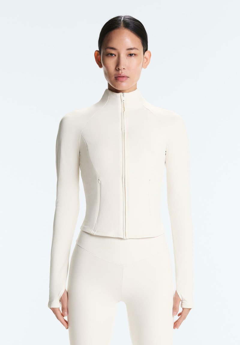 Femme portant un haut ajusté crème à manches longues avec fermeture éclair et trous pour les pouces ainsi qu’un legging assorti taille haute, debout devant un fond uni.