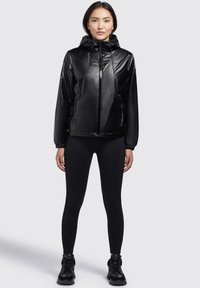 Chaqueta impermeable negra con acabado brillante, cierre delantero de cremallera y capucha con cordón. Presenta puños elásticos y dos bolsillos laterales.