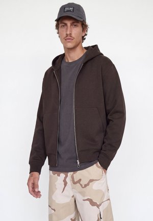 Homme portant un sweat à capuche noir zippé, un t-shirt gris foncé, un pantalon camouflage beige et marron, et une casquette noire avec un logo blanc.
