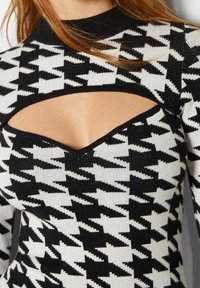 Svart och vit houndstooth-mönstrad stickad topp med hög krage, med en utskärning nedanför kragen och en figurnära design.