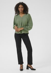 Olijfgroene blouse met lange mouwen, een v-halslijn en pofmouwen; gedragen met zwarte rechte broek en zwarte hakken.