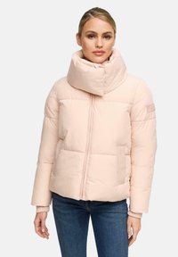 Frau trägt eine hellrosa Steppjacke mit hoch gepolstertem Kragen, durchgehendem Reißverschluss, Seitentaschen und blauen Jeans vor einem einfarbigen Hintergrund.