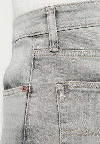 Τα γκρι τζιν από denim διαθέτουν κλασικό σχεδιασμό με πέντε τσέπες, ορατή ραφή, λεπτομέρειες με λογότυπο και ελαφριά φθαρμένη υφή κοντά στις τσέπες.